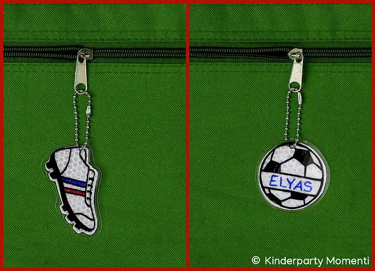 Zwei reflektierende Anhänger an grüner Stofftasche, einer in Form eines Fußballschuhs mit blauen und roten Streifen, der andere rund mit einem Fußballmuster und dem Namen 'ELYAS'