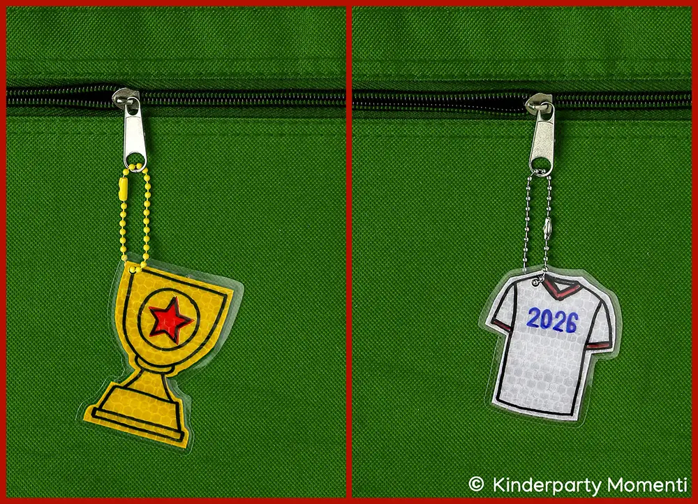 DIY Reflektor Anhänger - Fußballpokal & Fußball Trikot Zwei Schlüsselanhänger an Reißverschlüssen auf grünem Stoff, einer in Form eines Pokals mit rotem Stern, der andere in Form eines Trikots mit der Zahl '2026'