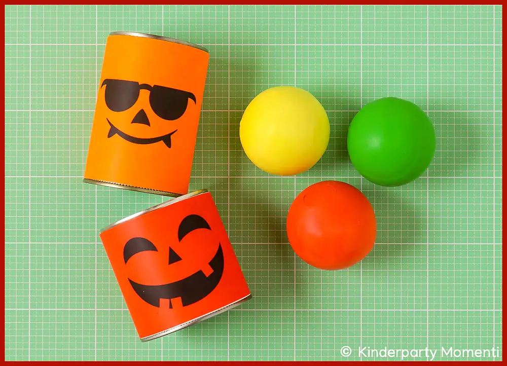 DIY Material für Dosenwerfen - zwei Dosen mit fröhlichen Halloween Kürbisgesichtern, daneben drei bunte Bälle auf grün kariertem Untergrund.