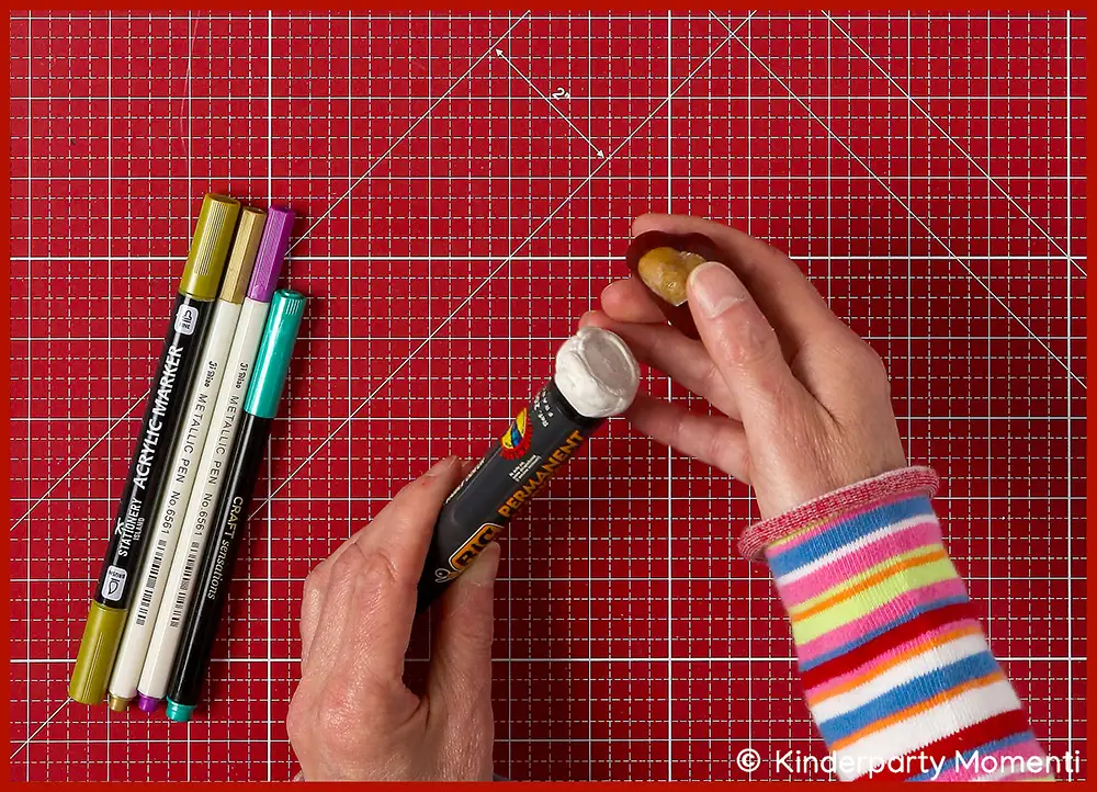 Hand hält Kastanie und schwarzen Permanentmarker, daneben liegen weitere bunte Marker auf einer roten Bastelmatte