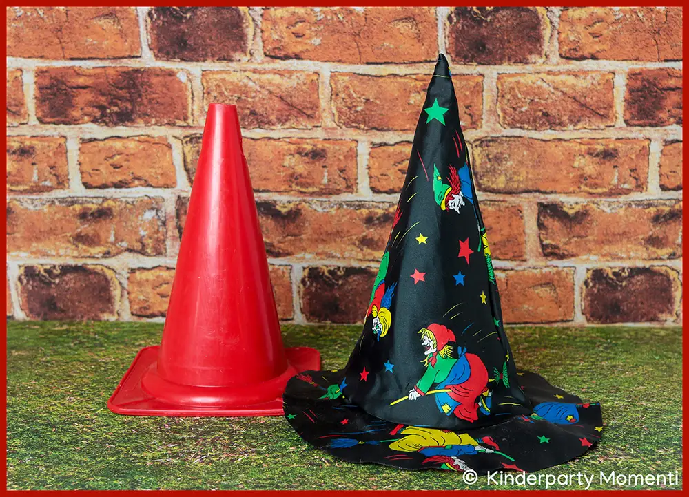Hexenhut als Pylone für Halloween Spiele mit Kindern Roter Verkehrskegel und daneben ein weiterer mit einem schwarzen Hexenhut mit bunten Sternen und Hexenmotiven verkleideter Pylone vor Ziegelsteinwand.