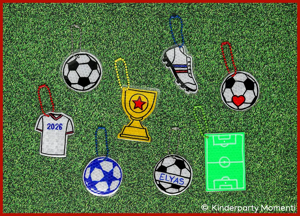 Selber gebastelte Fußball Anhänger aus Reflektorband Acht selbstgebastelte Fußball-Reflektoren in Form von Trikot, Pokal, Fußballschuh, Fußball mit Herz, Fußball mit Namen, Fußball mit Stern und Fußballfeld auf grünem Rasen mit bunten Ketten zum Anhängen