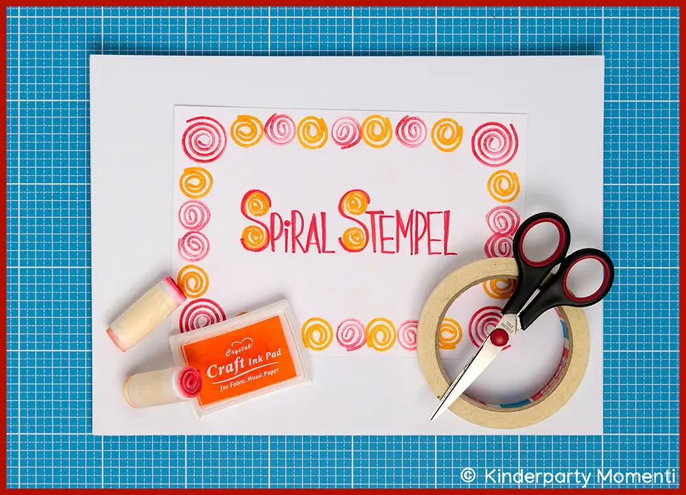 Blatt mit roten und gelben Spiralen und der Aufschrift 'SPIRAL STEMPEL' umgeben von Bastelmaterialien wie Schere, Klebeband, Stempelkissen und zwei Holzstempeln