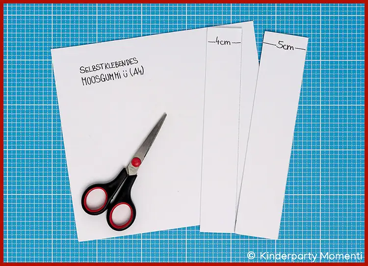 Schere liegt auf zugeschnittenen weißen Moosgummibögen mit handschriftlichen Maßen und Text auf blauem Schneideunterlage