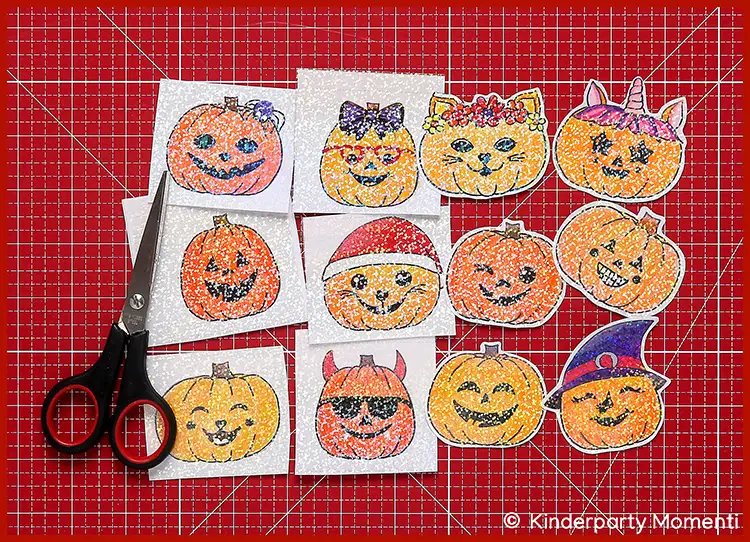 Holografische Sticker mit Kürbis Gesichtern ausschneiden Mehrere quadratische Aufkleber mit glitzernden Halloween-Motiven wie Kürbissen, Katzen und einem Teufel, daneben eine Schere auf einer roten Schneidematte