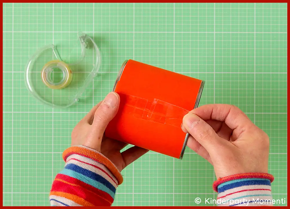 Hände kleben ein rotes Papier um eine Dose für Dosenwerfen Spiel,  grün karierter Untergrund, Klebebandspender liegt daneben.
