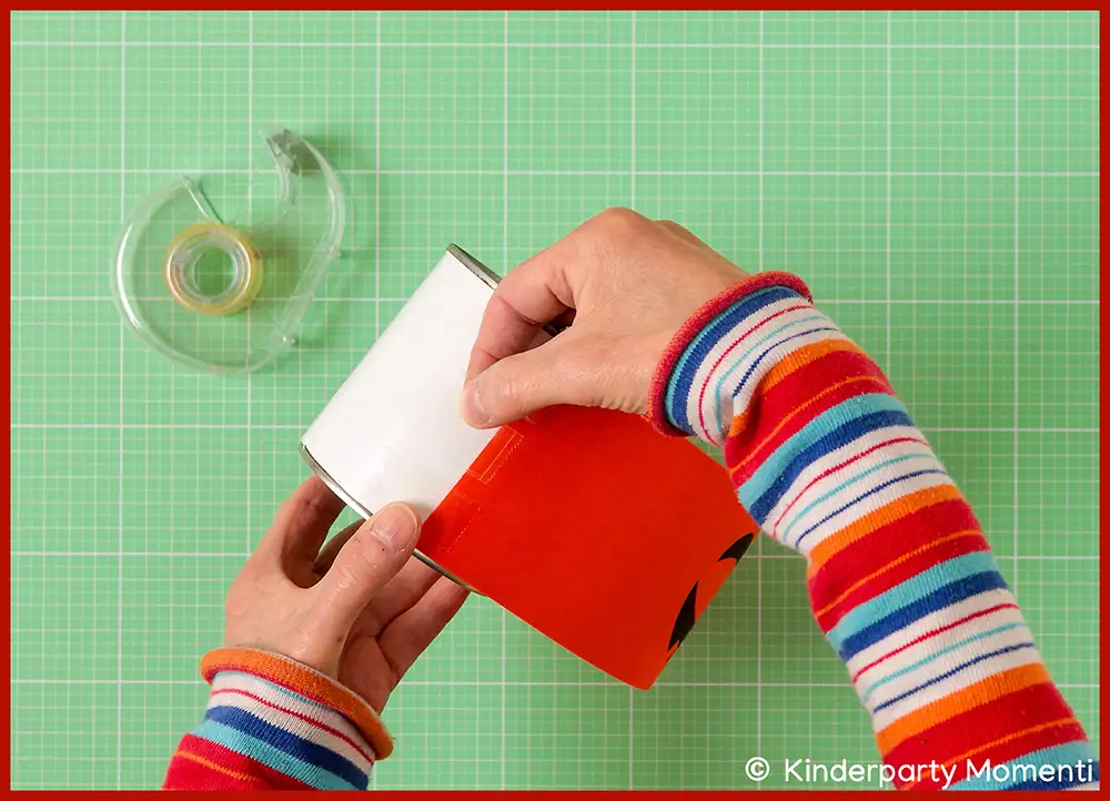 Hände bekleben eine leere Dose für Dosenwerfen mit rotem Papier auf grün kariertem Untergrund, daneben Klebeband