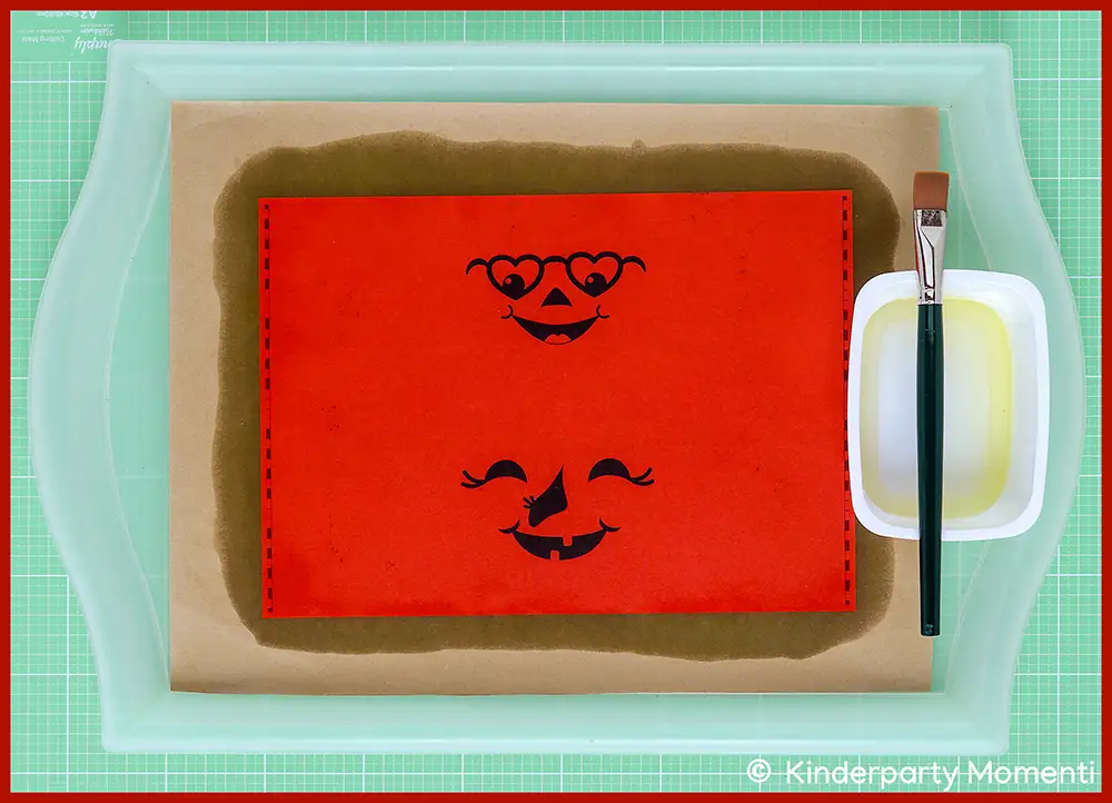 Rotes Papier mit zwei lachenden Kürbisgesichtern liegt auf braunem Papier in einem hellgrünen Tablett neben einem Pinsel und einer weißen Schale mit Öl