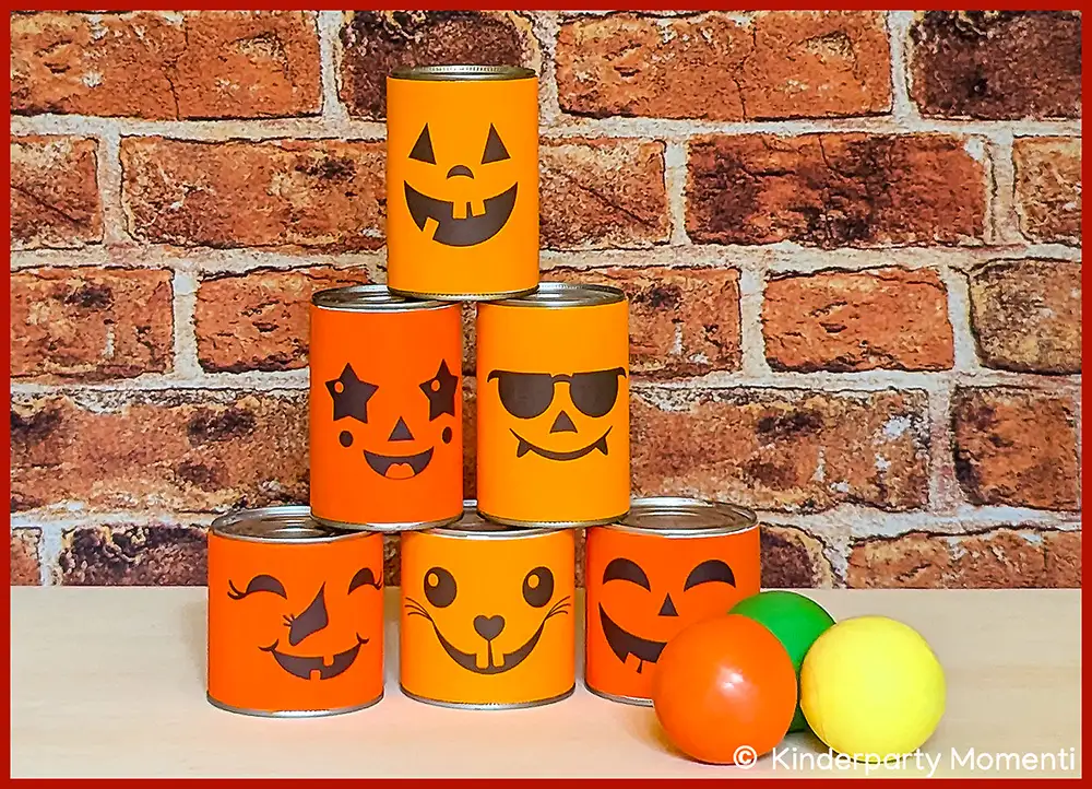 Sechs Dosen mit orangefarbenen Halloween-Gesichtern als Dosenwurfspiel vor roter Backsteinwand, daneben drei bunte Bälle
