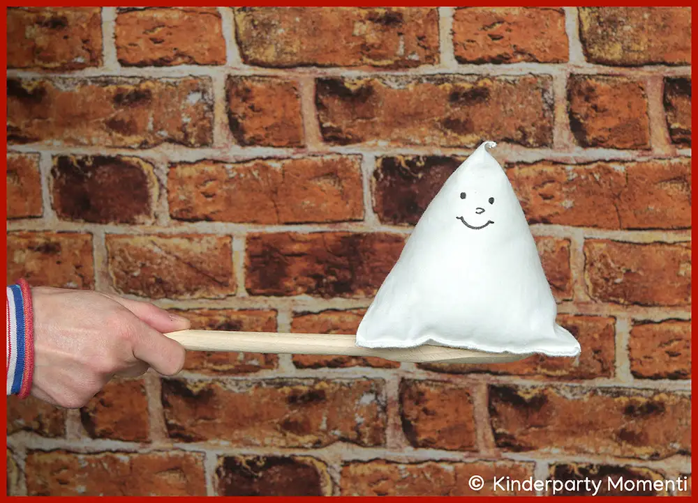 Kindergeburtstag Spiele für kleine Kinder - Eierlauf Spiel mit kleinen Stoffgespenstern Als Sinnbild für eine Eierlauf Spiel Variante hält eine Hand einen Holzlöffel mit weißem Stoff-Gespenst, das ein lächelndes Gesicht hat,