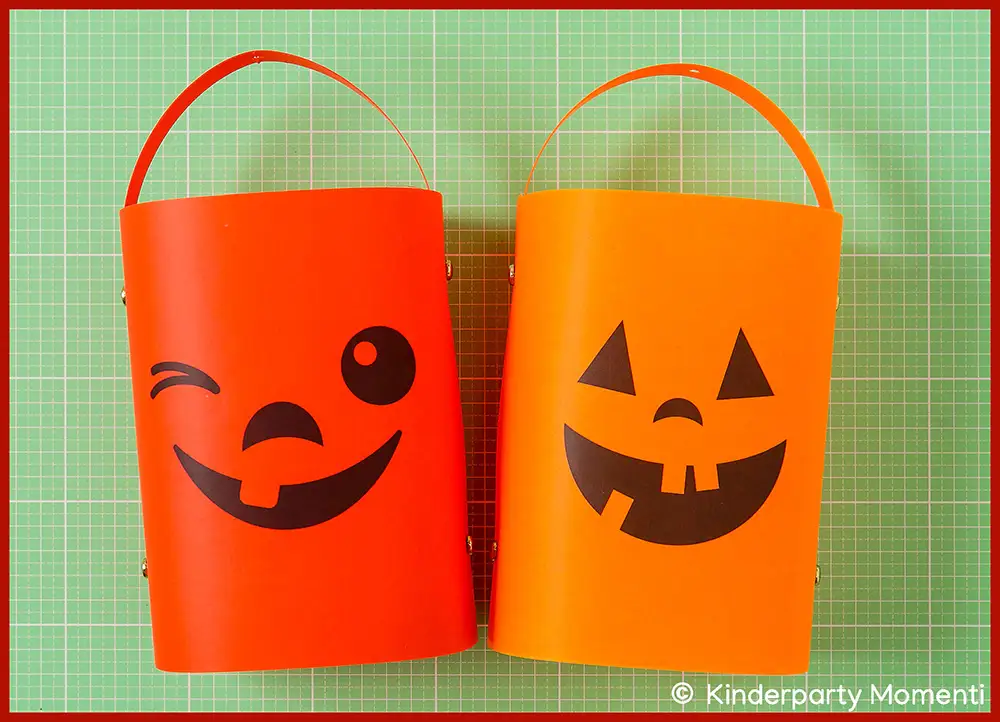 Zwei einfache Laternen aus orangefarbenem Papier mit schwarzen Kürbisgesichtern, eine zwinkert, die andere hat dreieckige Augen liegen auf einer grünen Bastelmatte
