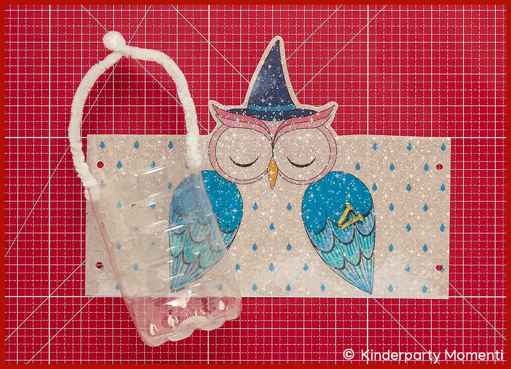 Mit blauen Farben ausgemalte Papier Vorlage für eine Eulen Laterne mit Hexenhut, davor eine transparente Plastikflasche mit weißem Griff, alles liegt auf einer roten Bastelmatte