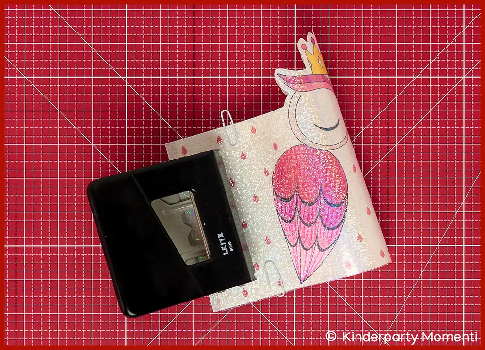 Rosa bemalte Eulen Laterne aus Papier wird mit schwarzem Locher gelocht, alles steht auf einer roten Bastelmatte