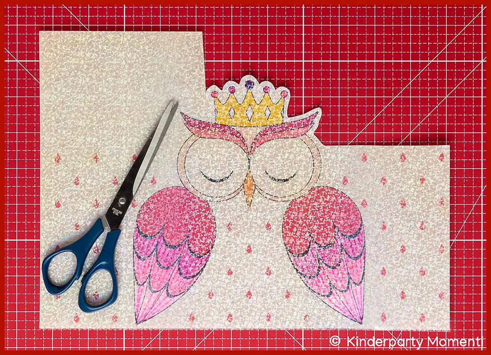 Mit rosa Farben ausgemalte Vorlage für eine Eulenlaterne, die eine Krone trägt, danben eine Schere, alles liegt auf einer roten Bastelmatte