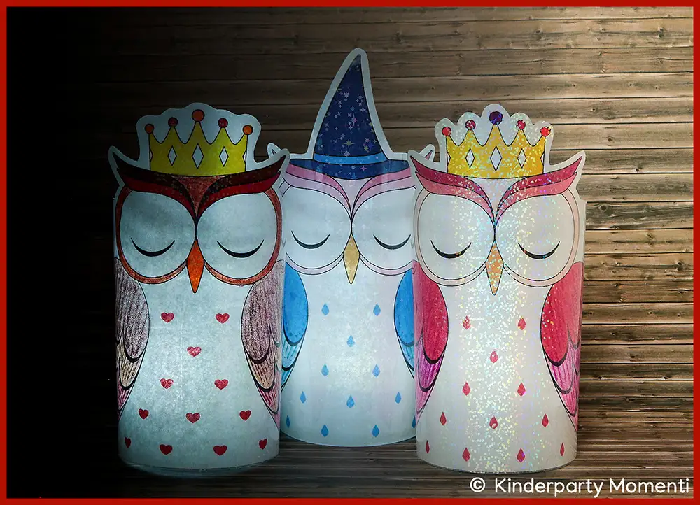 Drei leuchtende Eulen DIY Windlichter aus Papier, zwei mit goldener Krone und eine mit lila Hexenhut, vor Holzwand..