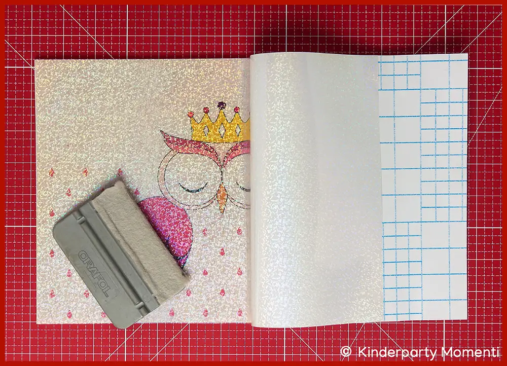 Bild zeigt wie rosa bemalte Eulen Laterne aus Papier mit glitzernder holographische Folie beklebt wird, die Eulenlaterne liegt auf einer roten Schneidematte und auf der Laterne ein grauer Rakel