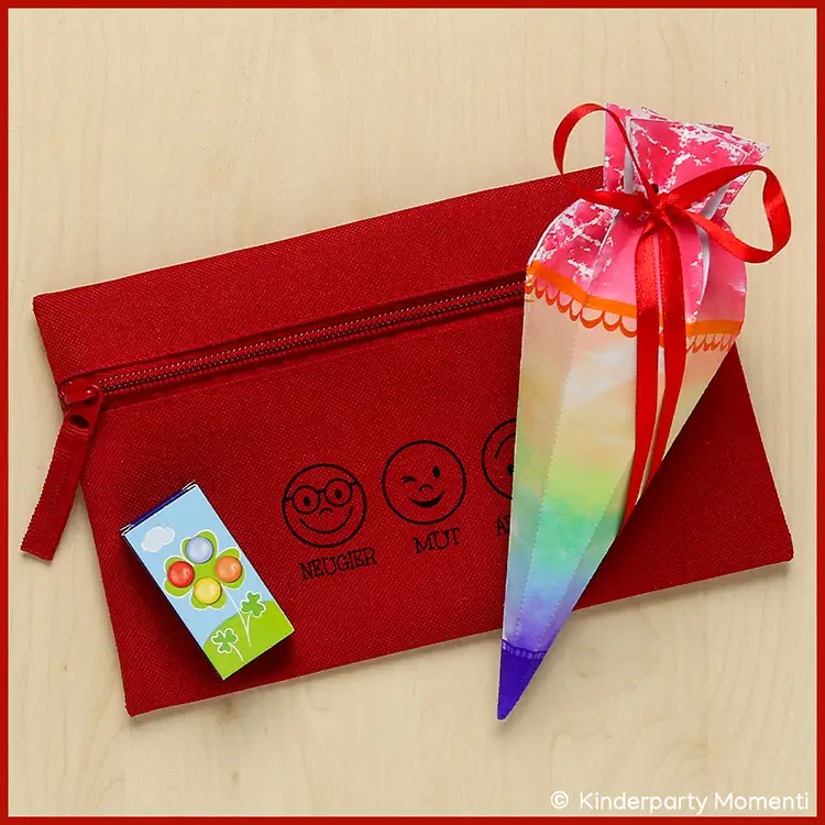 Rotes Federmäppchen mit drei aufgedruckten Emojis und den Worten 'NEUGIER', 'MUT', 'AUSDAUER', daneben eine bunte Schultüte mit rotem Band und ein kleines Päckchen mit Fruchtmotiven