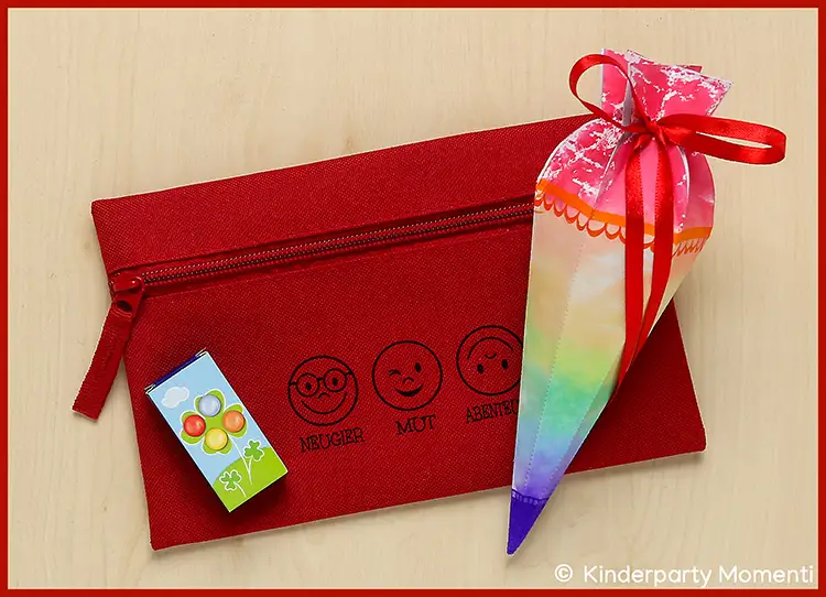 Rotes Federmäppchen mit drei aufgedruckten Gesichtern und den Worten Neugier, Mut, Abenteuer daneben eine kleine Schultüte mit Regenbogenmuster und roter Schleife sowie ein kleines Päckchen Süßigkeiten