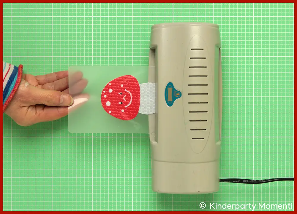 Hand schiebt Laminierfolie mit einem roten Fliegenpilz mit weißen Punkten in ein graues Laminiergerät auf grüner Bastelmatte