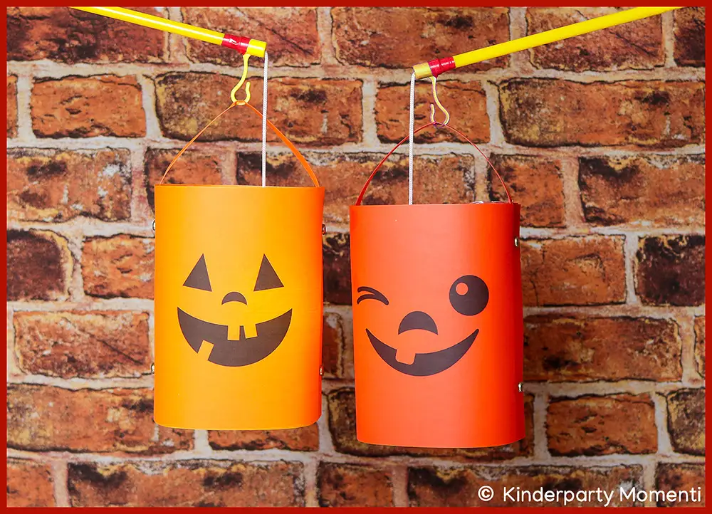 Zwei orangefarbene Halloween-Laternen mit lachenden Gesichtern hängen vor einer Ziegelsteinwand an gelben Stäben.