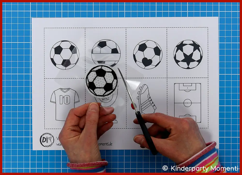 Bastelschritt 4 - Fußball Motive ausschneiden Hände schneiden mit einer Schere einen schwarz-weißen Fußball aus, im HIntergrund ein Blatt mit verschiedenen Fußballmotiven auf einem blauen Schneideuntergrund