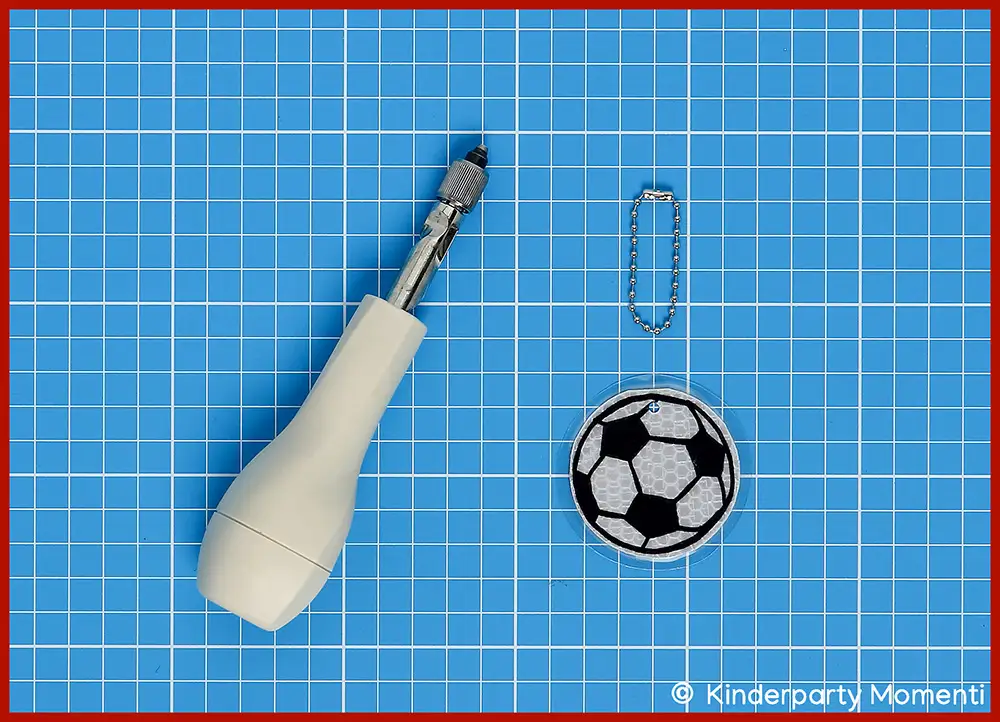 Bastelschritt 5 - Fußball Anhänger für Kugelkette lochen Fußball-Anhänger mit Kugelkette neben einem weißen Handbohrer auf Bastelmatte