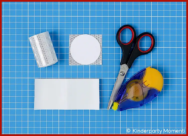 Auf blauer Schneidematte liegen eine Schere mit schwarzen Griffen, ein Kleberoller, ein rechteckiges und ein rundes Stück reflektierendes Material sowie eine Rolle reflektierendes Band