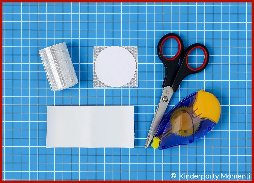 Bastelschritt 1 - Reflektorband für Fußball Taschenanhänger zuschneiden Auf blauer Schneidematte liegen eine Schere mit schwarzen Griffen, ein Kleberoller, ein rechteckiges und ein rundes Stück reflektierendes Material sowie eine Rolle reflektierendes Band