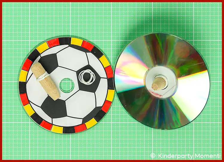 Zwei CDs auf grüner Bastelmatte, eine als Fußball bemalt mit Gummiband und Holzdübel, die andere blank mit Holzdübel in der Mitte