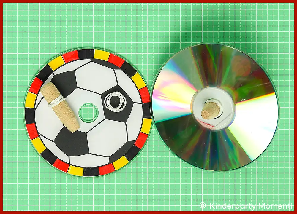 Holzdübel Kreiselpitze mit Gummis an CD befestigen Zwei CDs auf grüner Bastelmatte, eine als Fußball bemalt mit Gummiband und Holzdübel, die andere blank mit Holzdübel in der Mitte