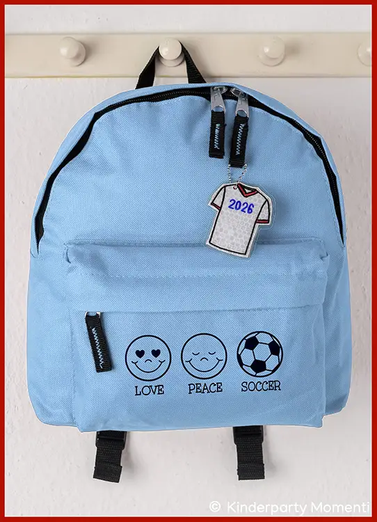 Hellblauer Kindergartenrucksack mit drei schwarzen Symbolen und Texten darunter: Herzaugen-Smiley mit 'Love', lächelndes Gesicht mit 'Peace' und Fußball mit 'Soccer', dazu ein Reflektor Taschenanhänger in Form eines Trikots mit der Zahl '2026'