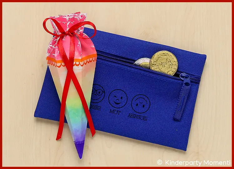 Feder-Mäppchen als Geschenk zur Einschulung Blaues Mäppchen mit Reißverschluss und drei aufgedruckten Emojis neben einer bunten Schultüte mit roter Schleife