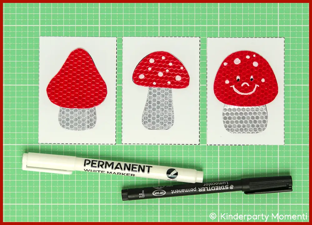 Drei rote Fliegenpilze mit weißen Punkten, einer mit lachendem Gesicht, darunter zwei Marker, weiß und schwarz auf gruner Bastelmatte