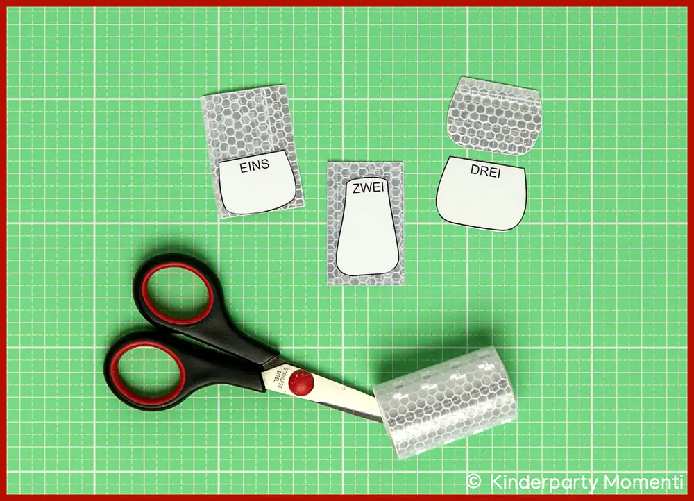 Schere mit schwarzen Griffen liegt auf grüner Bastelmatte, daneben drei ausgeschnittene kleine Pilz Stiel Vorlagen mit den Worten 'EINS', 'ZWEI' und 'DREI' auf Stücken von Reflektorfolie