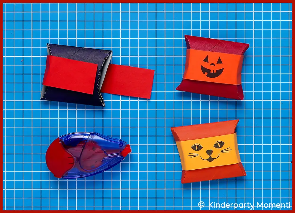 DIY Kissenschachteln mit Halloween Motiven basteln Drei kleine Geschenkboxen aus Toilettenpapierrollen mit Halloween-Motiven und Kleberoller, auf blauer Schneidematte