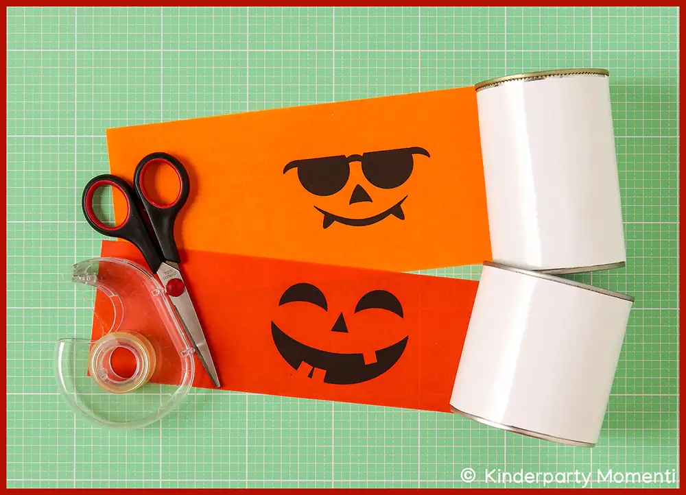 Zwei weiße Dosen, zwei Druckvorlagen mit schwarzen Halloween Kürbisgesichtern auf orangefarbenen Papier, daneben Schere und Klebeband auf grünem Schneideunterlage.
