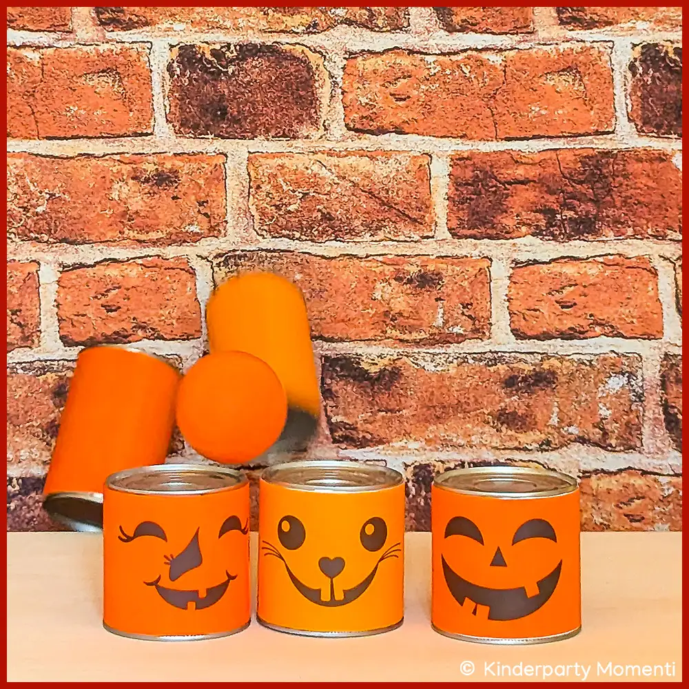 DIY Dosenwerfen für Halloween beim Wurf Dosen Wurfspiel - Drei Dosen mit orangefarbenen Halloween-Gesichtern vor Ziegelsteinwand, drei weitere Dosen fallen im Hintergrund.