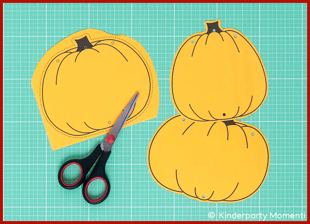 Kürbis Halloween Girlanden basteln mit Papier Auf einer Bastelmatte liegen drei gelbe Kürbisse aus Papier und eine Schere.