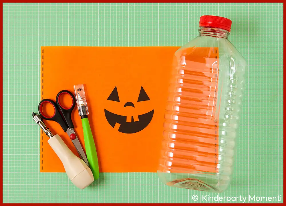 Laternen Vorlage aus orangefarbenem Papier mit Halloween Kürbisgesicht, daneben Schere, Klebestift, Stift und durchsichtige Plastikflasche mit rotem Deckel auf grüner Bastelmatte