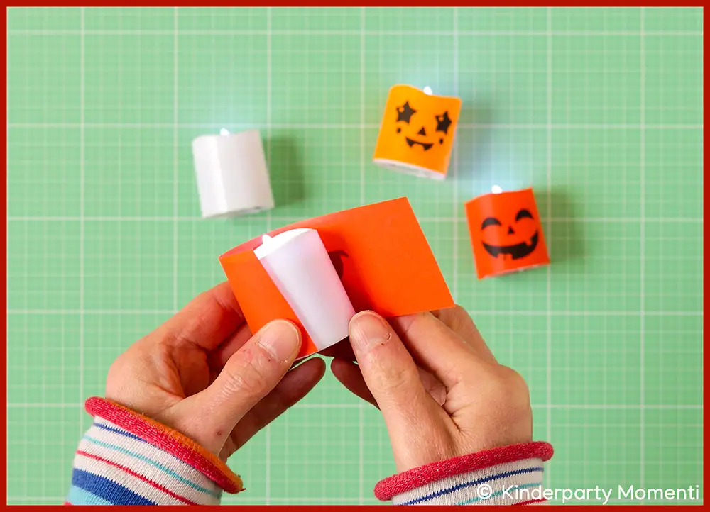 Hände kleben orangefabene Hülle um weißes LED Teelicht, weitere Teelichter mit Halloween Gesichtern liegen auf grüner Bastelmatte