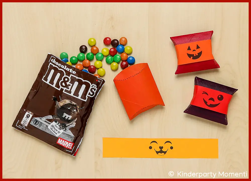 Kleine DIY Halloween Verpackungen aus leeren Toilettenpapier basteln Mehrere bunte Schokolinsen neben einer geöffneten Packung M&M'S, zwei kleinen Kissenschachteln mit Halloween-Gesichtern und einem orangen Papierstreifen mit einem lachenden Gesicht auf hellem Holztisch