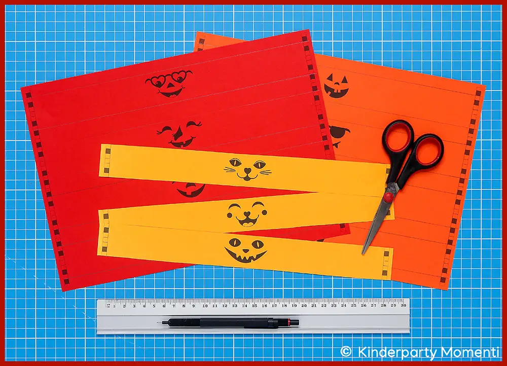 Halloween Vorlagen mit kleinen Kürbisgesichtern als Kissenschachtel Banderole Mehrere orange und gelbe Papierstreifen mit aufgedruckten Halloween-Gesichtern liegen auf blauer Schneidematte neben einer Schere, einem Lineal und einem Stift