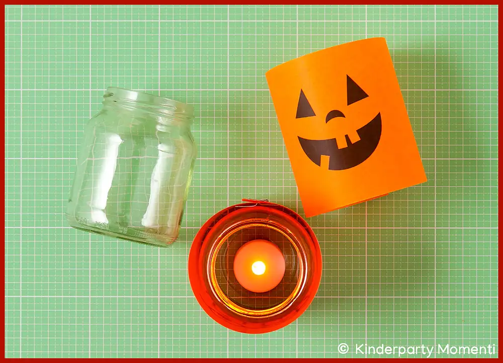 Leeres Glas neben orangefarbenem DIY Windlicht mit lachendem Kürbisgesicht und brennender LED-Kerze in rotem Glas auf grüner Bastelmatte