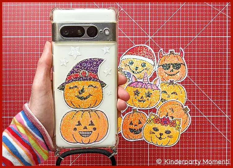 Glitzer Kürbis Sticker als Herbst Handy Deko Hand hält Smartphone mit durchsichtiger Hülle, darauf ein glitzernder Kürbissticker mit Hexenhut, daneben mehrere weitere glitzernde Kürbissticker mit verschiedenen Gesichtsausdrücken auf roter Schneideunterlage