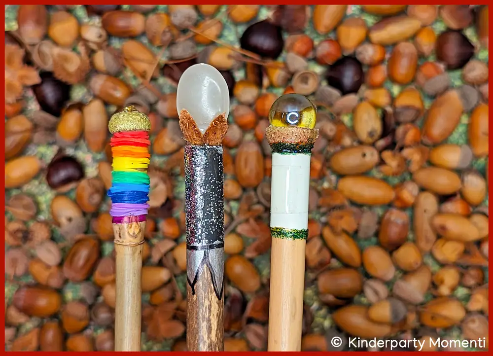 Hexen Zauberstab selbst machen im Herbst Drei bunte DIY Zauberstäbe mit verschiedenen Verzierungen, auf einem Hintergrund aus vielen Eicheln liegend.