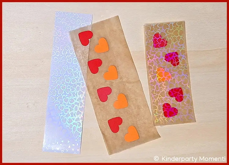 Herzen mit Holografischer Folie auf transparentes Klebeband kleben Drei längliche Aufkleber mit Herzmotiven, zwei mit holografischer Folie und einer mit roten und orangen Herzstickern auf Backpapier