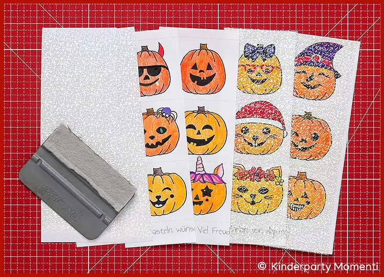 Selbst gemalte Sticker mit holografischer Folie bekleben Mehrere holografische Stickerbögen mit Halloween-Motiven wie Kürbissen und Katzen sowie ein grauer Rakel liegen auf einer roten Schneidematte