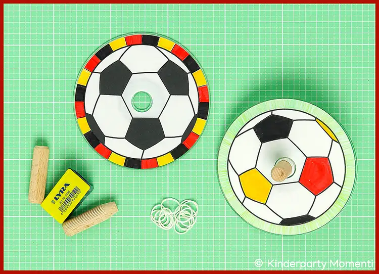 Zwei Kreisel mit schwarz-weißen Fußballmustern und farbigen Rändern, zwei Holzdübel und mehrere weiße Gummiringe auf grüner Bastelmatte