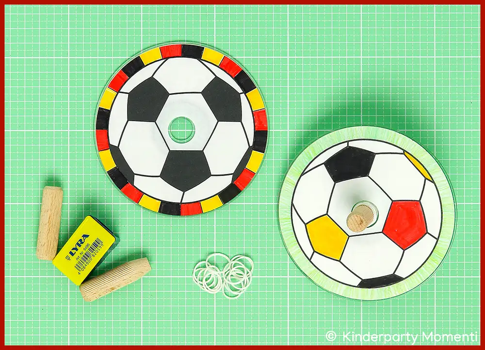Holzdübel als Kreiselspitze für Fußball CD Kreisel mit großem Anspitzer anspitzen Zwei Kreisel mit schwarz-weißen Fußballmustern und farbigen Rändern, zwei Holzdübel und mehrere weiße Gummiringe auf grüner Bastelmatte