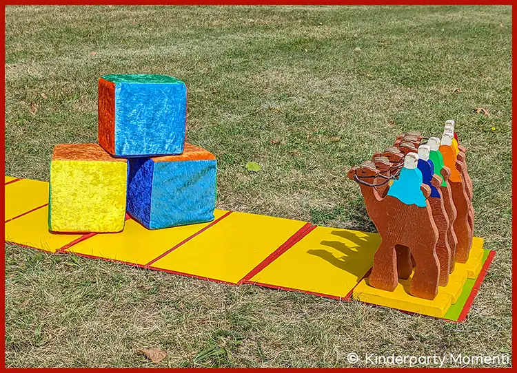 Kamelrennen Spielfeld für Outdoor Farbwürfelspiel Mehrere hölzerne Kamele in einer Reihe auf einem gelben Spielfeld neben drei großen bunten Würfeln auf Gras